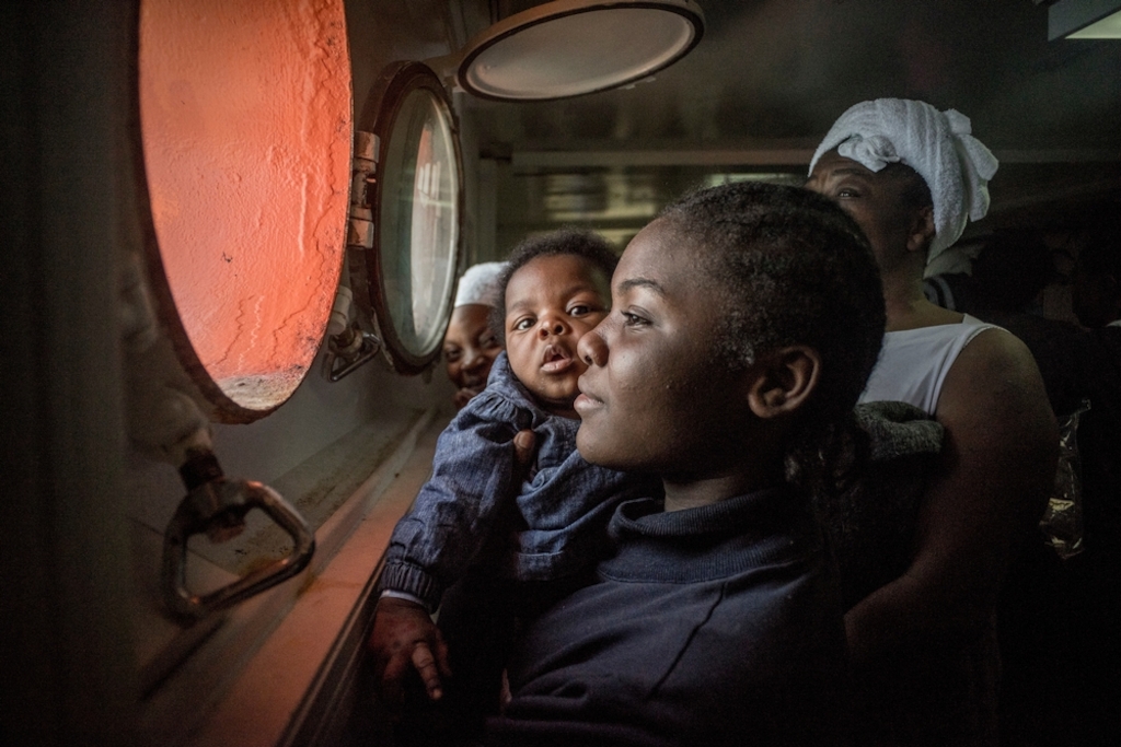 A woman and her baby look out of a porthole of an MSF search and rescue ship, run in cooperation with SOS Méditerranée, off the coast of Libya. Photographer: yann Merlin/SOS MEDITERRANEE